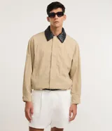 Campera de sarga color beige con corte oversized, cuello de cuero sintético negro con textura trenzada, cierre frontal oculto y bolsillos laterales.