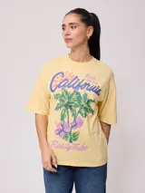 Remera color amarillo pastel con estampado de palmeras, flores de hibisco y texto 'California Ride the Tide Rusty Tribe'.