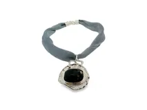 Collar con colgante de metal plateado con textura orgánica e irregular y piedra oscura en el centro, suspendido de un cordón de malla gris.