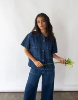 Camisa de denim azul de algodón reciclado, corte amplio, con cuello clásico, manga corta, cierre frontal con botones y dos bolsillos de parche en el pecho.