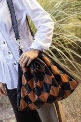 Bolso de hombro tipo hobo con diseño de rombos entrelazados en cuero negro y marrón. Presenta una silueta amplia y flexible con detalles de flecos largos en la parte superior.