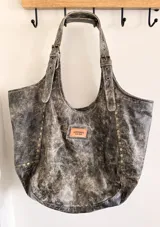 Cartera tote bag tipo shopper artesanal, elaborada en cuero con efecto desgastado color gris y negro. Cuenta con amplias asas de hombro regulables, tachas decorativas en la base y un parche frontal de cuero con el logo de la marca.