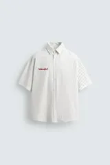 Camisa de algodón blanca con finas rayas verticales negras, corte relajado (relaxed fit) y manga corta. Presenta un bordado en hilo rojo en el lado izquierdo del pecho.