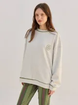 Buzo oversize color gris melange con corte crew, cuello redondo, mangas largas y detalles bordados en color verde en el cuello, puños y ruedo. Presenta el logo de la marca bordado en el pecho.
