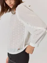 Camisa blanca de algodón con mangas largas abullonadas, detalle de volados en los hombros y puños elásticos. Presenta un diseño con cuello redondo y corte holgado.