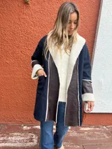 Saco de paño azul marino con solapas amplias y puños en corderito sintético color crema. Presenta detalles en cuero sintético marrón en la parte frontal y en las mangas, con un diseño abierto y corte recto.
