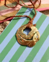 Collar artesanal con dije de flor en bronce o alpaca, suspendido en un cordón de seda natural teñido a mano. El diseño permite atarse con un moño, ofreciendo versatilidad para lucirlo de distintas formas.