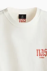Remera de algodón blanco crudo, de corte oversized con hombros caídos y cuello redondo acanalado. Presenta pequeño estampado en el pecho con la inscripción "nas ©1994" en tono anaranjado.