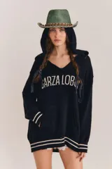 Sweater de punto negro con capucha y cuello en V, de manga larga y diseño holgado. Cuenta con el nombre de la marca bordado en el frente en color blanco, bolsillo estilo canguro, y detalles de rayas blancas en contraste en los puños y el ruedo.