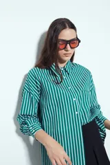 Camisa de silueta amplia y caída fluida, color celeste con rayas verticales blancas. Presenta cuello camisero clásico, cierre frontal con botones y un bolsillo aplicado en el pecho. Las costuras están resaltadas en azul oscuro.