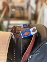 Cinturón de cuero en tono tostado con bordado a mano de hilos encerados en tonos marrones y beige con motivos geométricos, y hebilla metálica rectangular.
