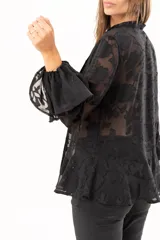 Blusa negra de tejido semitransparente con diseño floral en relieve, cuello alto con lazo frontal y mangas largas con volados en los puños.