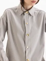 Camisa de algodón gris claro, de calce holgado y relajado, con cuello camisero clásico y mangas largas. Cuenta con botones metálicos dorados en forma de corazón en el frente.