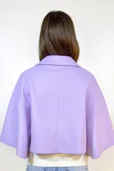 Capa corta de paño en color lila, con cuello redondo, cierre frontal de dos botones y mangas amplias tipo capa con pliegues laterales.