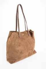 Bolso tipo tote de gran tamaño confeccionado en gamuza sintética color tostado. Presenta un diseño minimalista con asas largas de hombro y una pequeña placa metálica frontal con el logo de la marca.