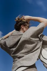 Capa amplia con capucha, confeccionada en lana fina sin teñir. Presenta cierre frontal, bolsillos delanteros y botones laterales que permiten ajustar la forma de las mangas.