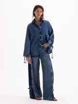 Camisa de denim azul profundo, de calce amplio y relajado, con cuello camisero clásico, hombros caídos y mangas largas con recortes y lazos regulables en los puños.