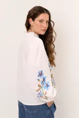 Camisa blanca de manga larga abullonada, con delicados bordados florales en tonos azules y verdes a lo largo de la botonadura frontal y en las mangas. Presenta cuello mao con reborde en contraste y cierre frontal con botones.