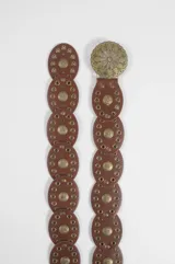 Cinturón de cuero vegano color marrón, compuesto por una serie de piezas circulares unidas, cada una con un aplique metálico central en forma de sol y detalles de tachas circulares. Incluye una hebilla metálica grande con diseño floral calado.