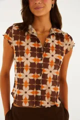 Top de manga corta con cuello camisero y cierre frontal de botones, confeccionado en tejido liviano con estampado geométrico de flores en tonos marrón, naranja y beige.
