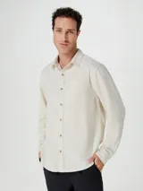 Camisa de manga larga en color beige, confeccionada en tejido de lino. Presenta cuello clásico, cierre frontal con botones a tono y un bolsillo aplicado en el pecho.