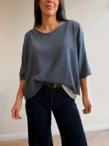 Conjunto de dos piezas compuesto por una remera básica blanca de algodón y un buzo liviano de textura plisada en color azul grisáceo. El buzo presenta un diseño de manga murciélago, corte holgado y detalles de hilo plateado en los bordes. Incluye un collar largo con dije circular de madera y metal.