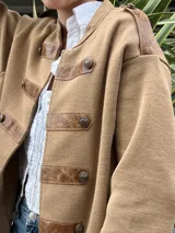 Chaqueta de abrigo color tostado, estilo militar, con detalles de apliques en cuerina marrón en los hombros, el cuello y las presillas frontales con botones metálicos dorados.