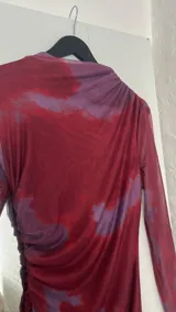 Vestido midi de microtul con diseño drapeado lateral, cuello alto y mangas largas. Presenta un estampado tipo tie-dye en tonos bordó y violeta.