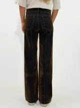 Pantalon de corte recto y tiro medio, confeccionado en denim con acabado aterciopelado en tono marrón oscuro. Presenta costuras verticales marcadas en contraste y cierre frontal con botón y cremallera.