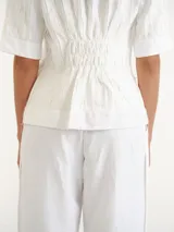 Blusa blanca de manga corta y cuello redondo, con diseño de rayas verticales texturizadas y detalles sutiles de flecos.