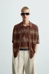 Camisa de punto de manga corta, color marrón rojizo con rayas horizontales en tono beige/verde oliva. Presenta cuello solapa y cierre frontal de botones.