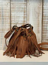 Cartera tipo bucket bag color marrón, confeccionada en cuero con textura granulada. Presenta un diseño con flecos largos en la parte frontal, herrajes metálicos en tono bronce y cierre mediante cordón ajustable. Incluye una correa larga regulable.