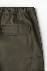 Pantalón cargo verde oliva de corte holgado, con cintura elástica ajustable con cordón, bolsillos laterales, bolsillos cargo con solapa y botón a presión en las piernas, bolsillos traseros con solapa y botón a presión, pliegues cosidos en las rodillas y cordón ajustable oculto en el bajo.
