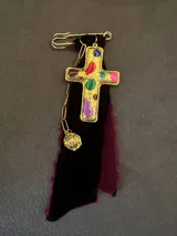 Pin decorativo con forma de cruz dorada de estilo vintage, adornada con piedras incrustadas. La pieza incluye una base de tela de terciopelo desflecada, una cadena metálica y un pequeño dije colgante.