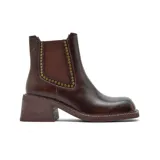 Bota tipo chelsea de cuero color marrón oscuro con acabado desgastado. Presenta paneles elásticos laterales decorados con tachas metálicas doradas, puntera cuadrada y taco bloque de altura media con textura amaderada.