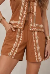 Conjunto de dos piezas color tostado con bordados florales en hilo blanco, compuesto por una blusa sin mangas con escote en V y cierre frontal con lazos, y un short tiro alto.