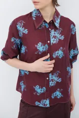 Camisa de corte clásico con cuello camisero y cierre frontal de botones, confeccionada en tela color bordó con estampado floral en tonos celestes.