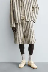 Bermuda de hombre color beige con estampado de rayas verticales azules con efecto desgastado. Corte relaxed fit, con cierre frontal de cremallera y botón, bolsillos delanteros y bolsillos traseros de plastrón.