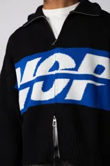 Campera tejida de punto con cierre frontal completo y cuello polo. Presenta un diseño con franja horizontal en color azul y letras en blanco sobre fondo negro, con puños y ruedo acanalados.