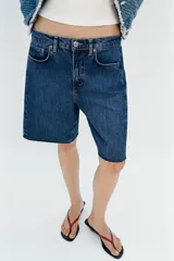 Bermuda de denim azul de tiro medio y corte relaxed, con diseño de cinco bolsillos y bajo desflecado. Cuenta con cierre frontal de cremallera y botón metálico.