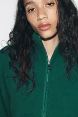 Jersey de punto de color beige con cuello subido y cierre frontal con cremallera metálica. Presenta mangas largas con puños acanalados y diseño de corte holgado.