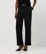 Pantalón de vestir negro confeccionado en malla punto roma, de corte largo y ligeramente afunilado. Presenta cintura alta con un adorno metálico decorativo en el frente y costuras verticales que estilizan la silueta.