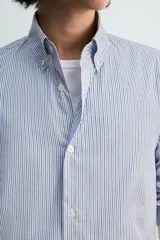 Camisa de algodón de corte relajado, con cuello solapa abotonado (button-down) y manga larga terminada en puño con botón. Presenta un patrón de rayas verticales finas en tonos azul claro y blanco.