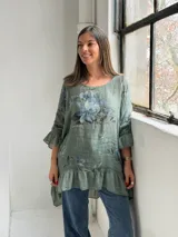 Blusa verde de lino con estampado floral en tonos azules y grises, escote redondo, mangas 3/4 con volados y ruedo asimétrico con volado fruncido.