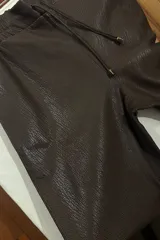Pantalón de cuero color chocolate con corte recto y holgado. Presenta cintura elástica con cordón ajustable y costuras horizontales en la parte inferior de las piernas.