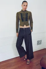 Chaqueta corta de antelina color verde militar, con cuello subido, manga larga y bajo asimétrico. Presenta detalles de alamares negros bordados en contraste en el frente y en los puños.