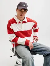 Buzo tipo polo de manga larga con diseño de franjas horizontales anchas en color rojo y blanco, cuello camisero blanco y cierre frontal parcial con solapa y botones ocultos.