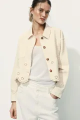 Cazadora color rosa pálido, de cuello solapa y manga larga. Tiene bordados perforados a tono en el ruedo y cierre frontal con botones color madera.