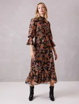 Vestido midi de gasa con estampado de arabescos en tonos cálidos sobre fondo negro. Presenta cuello alto con detalle de punto smock, mangas largas con puños elásticos y volados, y falda con tajo lateral y ruedo con volado. Incluye forro interno y cierre posterior.