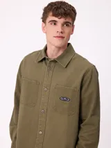 Camisa de gabardina color verde oliva, con cuello clásico, cierre frontal de botones y dos bolsillos frontales con parche. Incluye un pequeño parche con el logo de la marca en el bolsillo izquierdo.